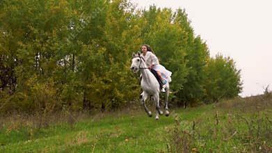 Long-haired woman in white dress is riding horse along wood