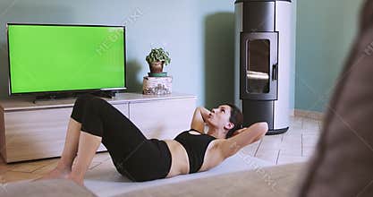 Woman exercising at home following a remote fitness class. Woman doing home training with green screen TV. Abs workout