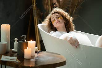 A young woman with eye patches lies in a bubble bath. A long-haired woman soaks in a bubble bath and does a wellness