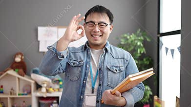 Young chinese man teacher holding books doing ok gesture at kindergarten