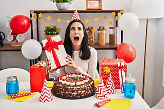 Hispanic woman celebrating birthday with cake holding present angry and mad screaming frustrated and furious, shouting with anger