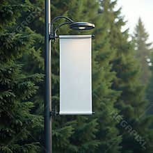 Blank vertical banner hangs from lamppost with blurred evergreen trees backdrop. White advertising board for message display.