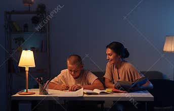 Teenage boy is carefully does his homework in notebook, mom holding a book, sits next to son and helping with assignment. Concept