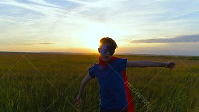 A boy in a superman costume runs across the green field at sunset
