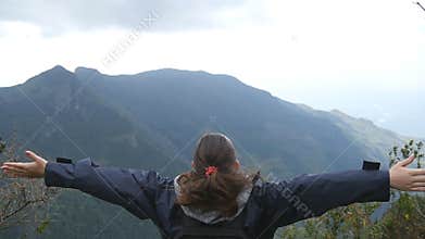 Young female hiker with backpack reaching up top of mountain and raised hands. Woman tourist standing on the edge of