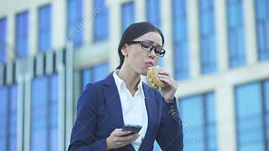 Female office manager eating unhealthy burger checking messages on phone, stress