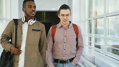 Two young handsome multi-ethnic friends walking down white glassy corridor in college talking passing by standing girls