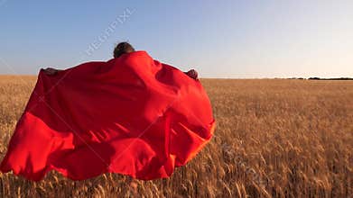 Girl super hero running across field with wheat in red cloak against blue sky.