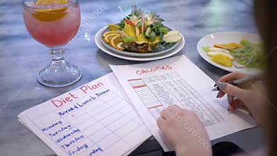 Nutritionist woman writes diet plan with food calorie sheet on table with punch and fruits in plate at cafe