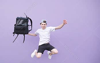 Positive young man in a white T-shirt jumping with a backpack against the background of a purple wall