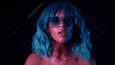 Portrait of unusual girl with blue hairstyle in neon light at black wall. Tempting woman with perfect makeup and wig