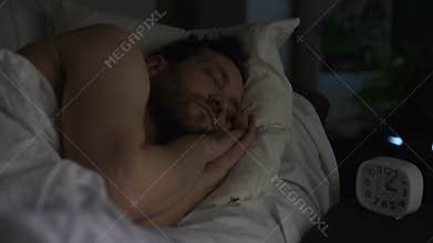 Bearded man sleeping on sofa bed, clock standing on night table, late night rest