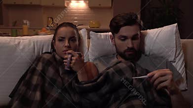 Sick couple sits together. They are covered with blanket. Girl drinks from cup. Guy get thermometer and look at it. He