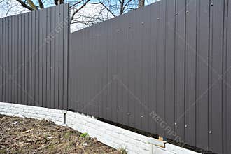 A close-up of a perimeter house fence made from metal corrugated gray fence sheets installed on a brick foundation that is turning