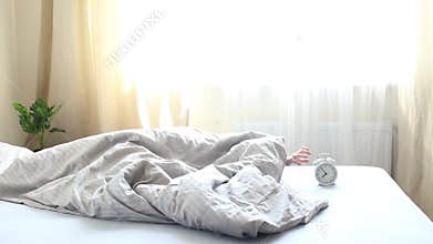 The child does not want to get out of bed and hides the alarm clock under the blanket.
