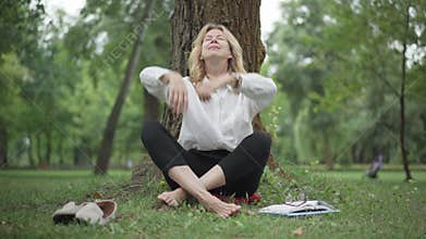 Portrait of confident middle aged barefoot businesswoman taking off eyeglasses and sitting in lotus pose in summer park