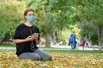 Young girl in protective mask sits on lawn in autumn park and listen to music