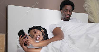 Suspicious young american dark skinned black husband laying in bed with her wife watching his girlfriend. Black girl