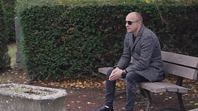 A stylish man in a gray overcoat and sunglasses is relaxing on a park bench, looking contemplative