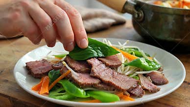 Hand garnishing sliced beef salad with fresh basil leaves on a white plate with vegetables for a healthy and delicious meal prep