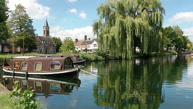 Scenic river view with houseboats willow tree and historic buildings under a bright blue sky reflecting in the calm water