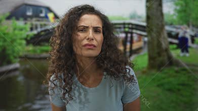 Woman frowns with furrowed brow in green park by a wooden bridge over a pond under soft daylight; quiet introspection