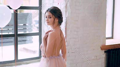 Lovely attractive girl in loose light dress with open bare back, standing at huge window, smiling tenderly, mysterious