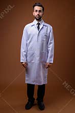 Young handsome bearded Iranian man doctor against brown backgrou