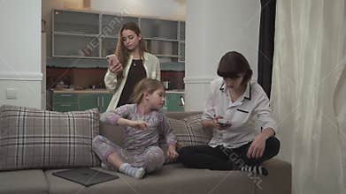 Two older sisters texting messages on cell phones younger girl typing on laptop sitting on the couch at home. Little