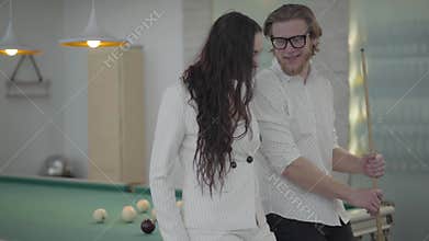 Portrait blond bearded man and his girlfriend talking near the billiard table. Confident man in white shirt explains to