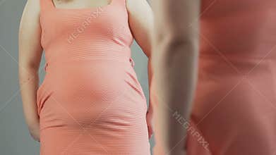 Woman pulling back dress and looking at her flabby stomach reflected in mirror