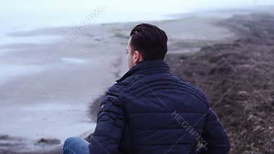 Pensive man sits on bare beach stones at vast grey sea