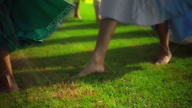 Legs of young European girls in long dresses walking in a circle with barefoot legs on a grass