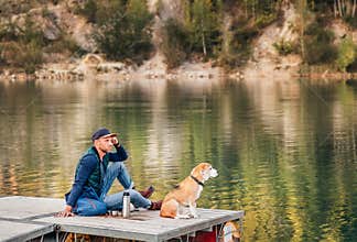 Man dog owner and his beagle friend sitting on the wooden pier on the mountain lake and enjoying the landscape during their