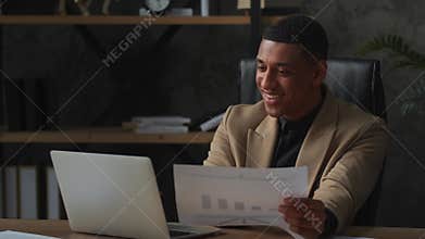 A smiling man in a business suit is sitting at a table and typing on a keyboard. Black male businessman talking to