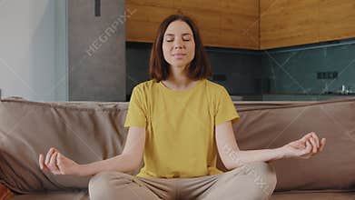 A young woman in a yellow t-shirt is captured meditating on a comfortable sofa in a modern living room setting. She sits