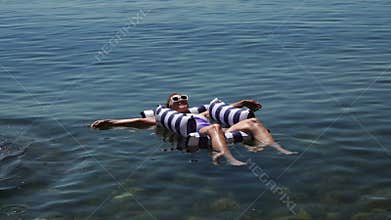 Woman Floating Water: Adult woman happily floats on striped raft in clear blue lake on a warm summer day for relaxation.