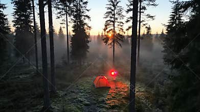 Sunrise Over Misty Forest Clearing With Orange Tent and Beacon Light in Remote Wilderness Landscape