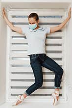A female ballerina in a mask in casual clothes in jeans and a t-shirt in Pointe shoes at home on the windowsill posing for the