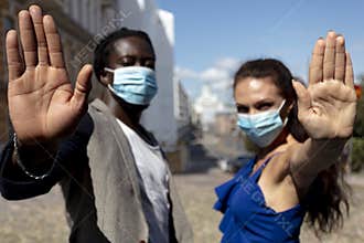 Portrait of a young European woman and African man wearing a mask in a city street. Stop coronavirus hand gesture