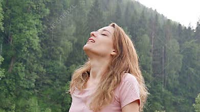 Beautiful happy girl standing in the summer rain against the backdrop of mountains covered with forests. Young woman