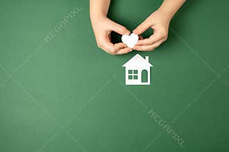 Family home and real estate concept. Close up of child hands holding white paper house and heart on green background.