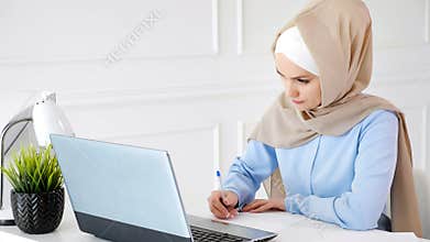 Young muslim woman student in hijab is doing her homework writing and drawing table on paper using laptop.
