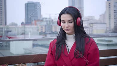 Portrait of confident Caucasian girl in red coat and gloves putting on headphones as sitting in city under light rain or