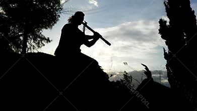 Native American flute player silhouette on a hill with trees in a cloudy sky background.
