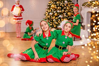 Two cheerful happy girls-friends in costumes of Santa`s elves on the background of a Christmas tree and other children