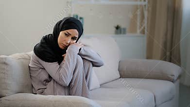 Anxious sad young woman in black hijab sitting on couch in living room suffering from psychological problem trouble at