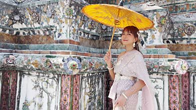 Young woman wearing Thai dress with accessories stands holding antique umbrella and bag walking at Wat Arun Ratchawararam