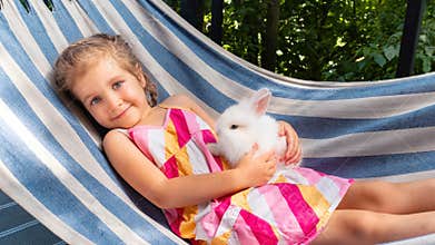 A girl with a white bunny lies on a hammock in the summer on a sunny day. White fluffy decorative rabbit in the arms of a little