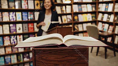Large open book displayed on a wooden pedestal in a bookstore, with an author in the background giving signed copies to